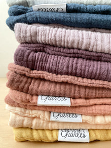 Stack of Charley Charles Burp Cloths in mustard, pale peach, dusty blush, nutmeg, plum, dusty purple, aegean blue, and light blue. 