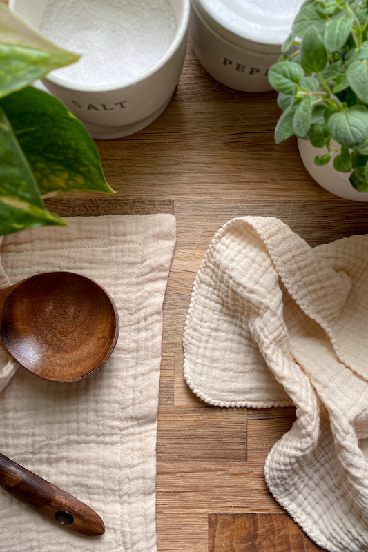 Two Charley Charles brand towels in the color Ecru. Thick 4 layer towel on the left with a wooden spoon laying on it. Thin 2 layer towel on the right. Towels are surrounded by plants and salt and pepper holders. 
